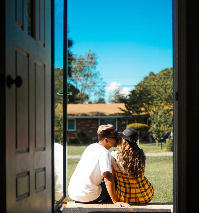 Couple kissing on a porch, seen through an open door, with a house and blue sky in the background.