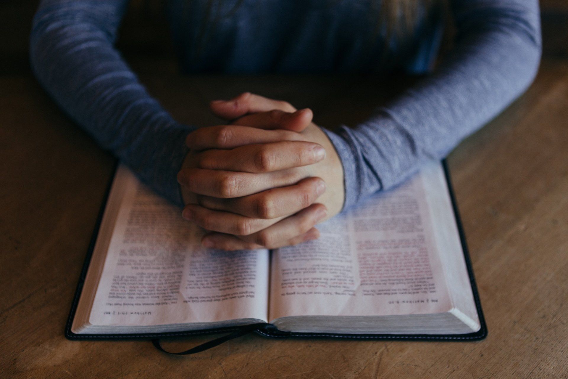 Hands clasped on top of an open book, likely a Bible, on a wooden surface.